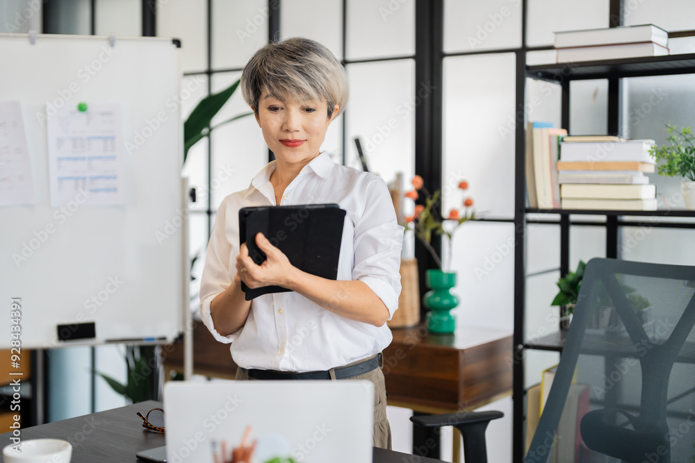 A middle-aged businesswoman with short gray hair is standing and working with a tablet in a modern office, surrounded by stylish decor and greenery A middle-aged businesswoman with short gray hair is standing and working with a tablet in a modern office, surrounded by stylish decor and greenery
