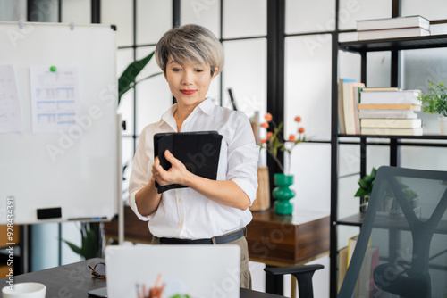 A middle-aged businesswoman with short gray hair is standing and working with a tablet in a modern office, surrounded by stylish decor and greenery