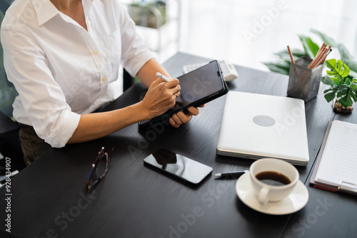 A middle-aged businesswoman is sitting at her desk in a modern office, working on a tablet with focus and concentration photo