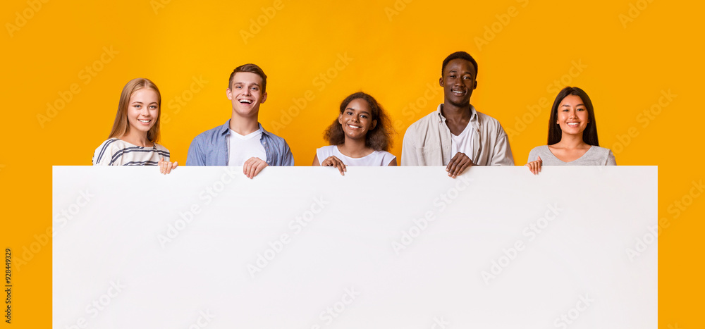 Smiling group of multiracial students standing together in row and displaying white empty placard, yellow studio background Smiling group of multiracial students standing together in row and displaying white empty placard, yellow studio background