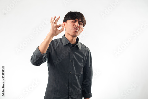 An Asian man in a gray shirt making an "OK" hand gesture with one hand while looking slightly upward, his lips pursed as if whistling. He stands against a plain white background