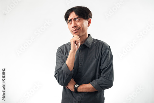 An Asian man in a gray shirt, standing with his hand on his chin, looking thoughtful or pondering. He has a slightly tilted head and a curious expression, against a plain white background.