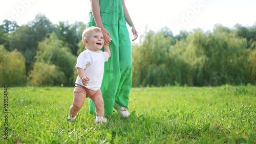 Mom and baby taking the first steps. Walk kid boy concept. Mom holds baby hand while walking along the bridge in the park. Mom holding kid hand walking lifestyle in summer.