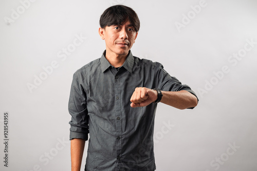 An Asian man in a gray shirt checking the time on his wristwatch, with a focused expression. The background is plain white, emphasizing his attention to the time