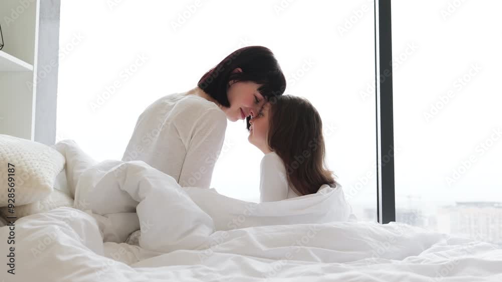 Back view of loving mother cuddling with her daughter in the morning. Cozy bedroom setting with soft natural light. Displays warmth, affection, family bond, and tenderness.
