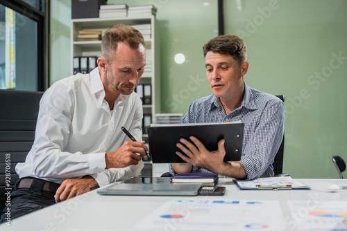 Caucasian middle-aged male businessperson and an Italian accountant are seated at a desk, engaging in a professional meeting, discussing various aspects of technology and innovation in their fields.