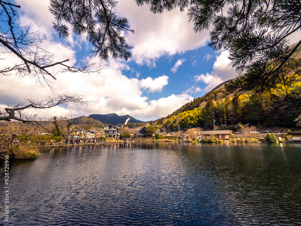Fototapeta premium the serene beauty of Kinrin Lake, framed by branches with a scenic mountain backdrop. The tranquil waters reflect the sky, while visitors enjoy the picturesque surroundings.