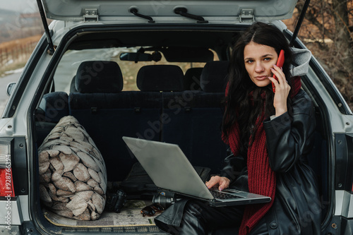 A young freelance woman is sitting on the trunk of her SUV and working on her laptop.