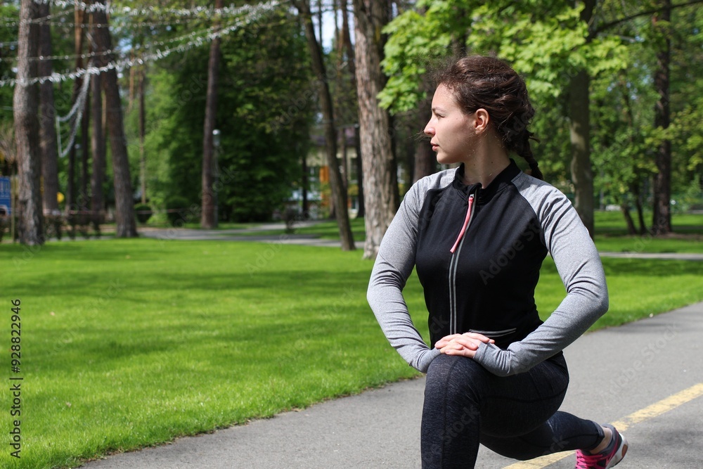 Young brunette athletic woman in grey sportswear stretching legs before running jogging in green park outdoor in sunny day.Concentrated millennial fitness girl doing lunges exercise,workout,training Young brunette athletic woman in grey sportswear stretching legs before running jogging in green park outdoor in sunny day.Concentrated millennial fitness girl doing lunges exercise,workout,training