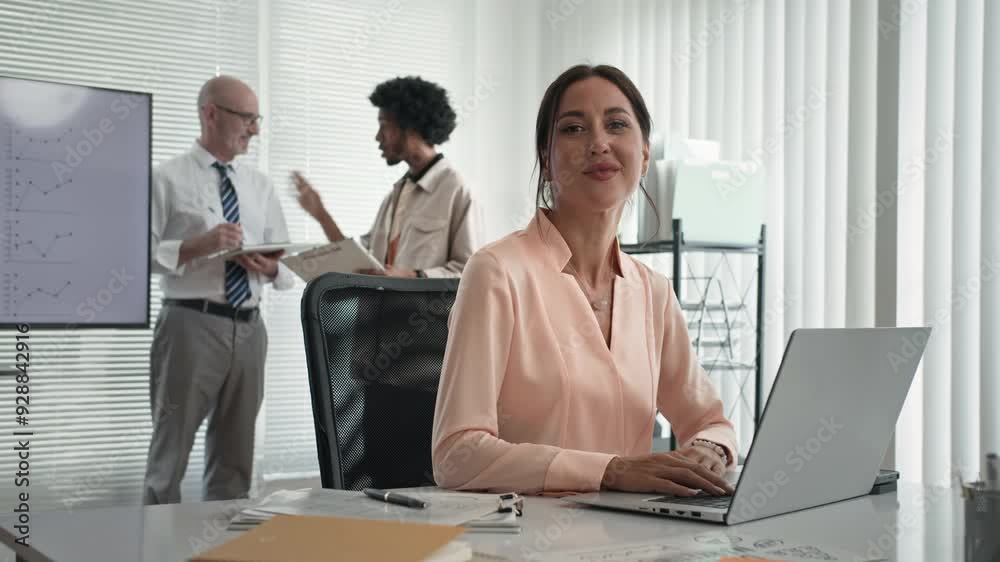 Portrait of young Caucasian female lawyer looking at camera when sitting at workplace and male colleagues communicating in background Portrait of young Caucasian female lawyer looking at camera when sitting at workplace and male colleagues communicating in background