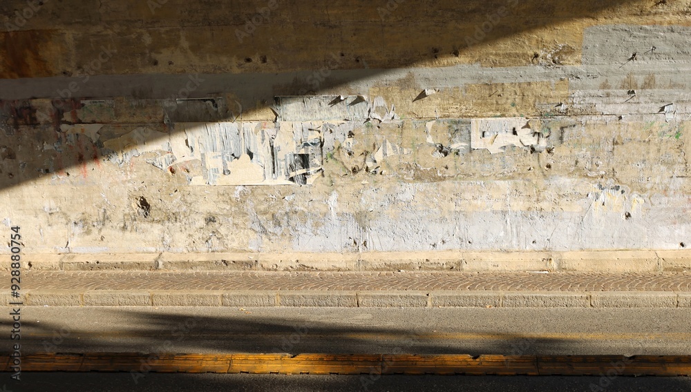 Street tunnel divided in two by the sunlight. Erased graffiti and ripped poster on the wall. Sidewalk and road in front. Grunge background for copy space. Street tunnel divided in two by the sunlight. Erased graffiti and ripped poster on the wall. Sidewalk and road in front. Grunge background for copy space.