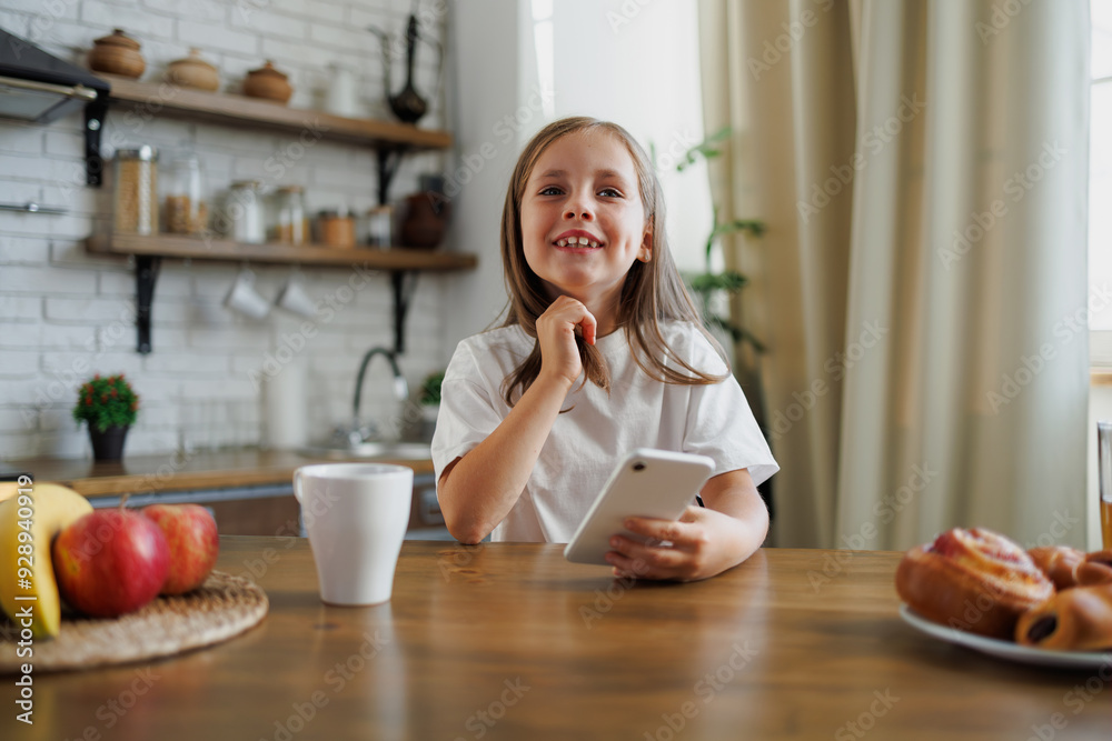 Positive kid holding cellphone near buns and fruits in kitchen Positive kid holding cellphone near buns and fruits in kitchen
