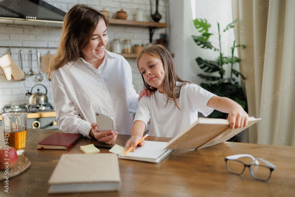 Smiling mother holding smartphone near daughter with books in kitchen Smiling mother holding smartphone near daughter with books in kitchen