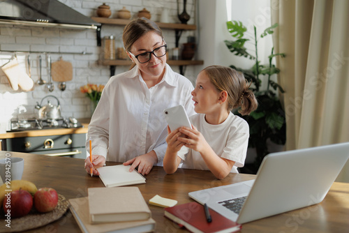 Child using smartphone near mother with notebook and laptop in kitchen