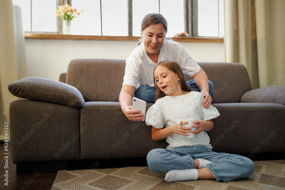 Smiling woman holding smartphone near daughter with cup at home Smiling woman holding smartphone near daughter with cup at home