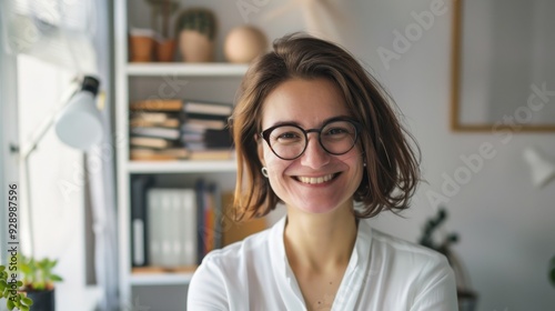 A smiling woman in glasses and a white shirt stands in a bright, organized home office, exuding joy and enthusiasm as she faces new challenges in her workspace.