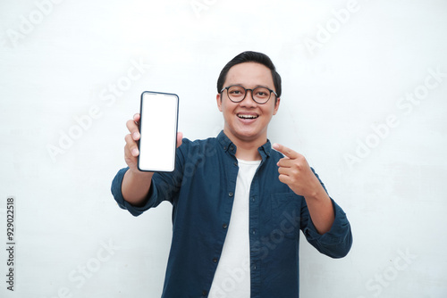 Happy Asian man holding blank smartphone screen with happy and excited expression. Young Asian man wearing blue shirt smiling and showing white mobile phone screen for advertisement.
