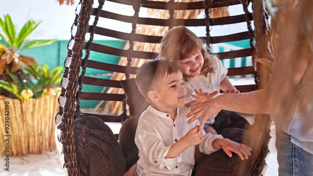 Two small children are sitting in suspended swing chair, their mother is standing next to them, they are having fun playing game showing symbols with hands. Concept of happy childhood and family. Two small children are sitting in suspended swing chair, their mother is standing next to them, they are having fun playing game showing symbols with hands. Concept of happy childhood and family.