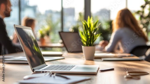 A laptop sits on a wooden table in an office setting with a plant, a phone, and two blurred people in the background.