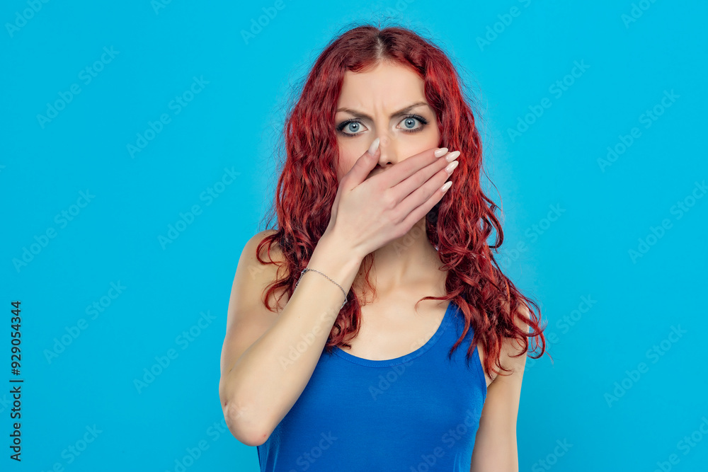 Portrait of a young angry woman in blue shirt, and long red wavy hair cover her mouth with a hand on a blue background Portrait of a young angry woman in blue shirt, and long red wavy hair cover her mouth with a hand on a blue background
