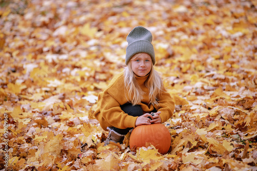 Child sits among golden autumn leaves in a park, holding a pumpkin, dressed in a cozy orange sweater and grey beanie, capturing a calm and seasonal moment. photo