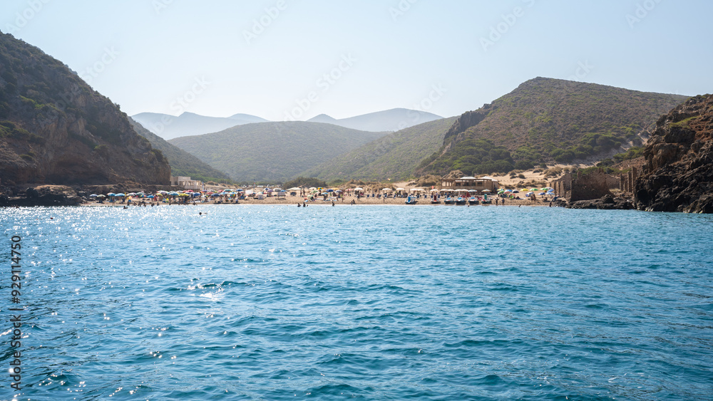 View from a boat of Cala Domestica, a unspoiled cove with with people sunbathing and hills in background in Sardinia island, Italy View from a boat of Cala Domestica, a unspoiled cove with with people sunbathing and hills in background in Sardinia island, Italy