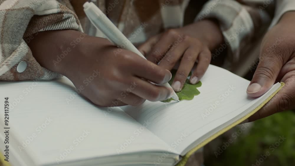 Hands of unrecognizable Black woman and junior child spending time outdoors holding notebook and tracing outline of green leaf using pen to create shape