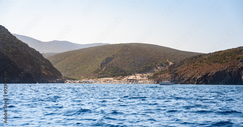 View of Cala Domestica from a boat, showcasing crystal-clear blue waters, rugged hills, and a serene beach with people sunbathing in the Sardinia's natural landscape, Italy View of Cala Domestica from a boat, showcasing crystal-clear blue waters, rugged hills, and a serene beach with people sunbathing in the Sardinia's natural landscape, Italy