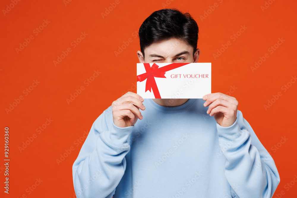 Young fun man he wearing blue sweatshirt casual clothes hold cover mouth with gift certificate coupon voucher card for store blink eye isolated on plain red orange color background. Lifestyle concept. Young fun man he wearing blue sweatshirt casual clothes hold cover mouth with gift certificate coupon voucher card for store blink eye isolated on plain red orange color background. Lifestyle concept.