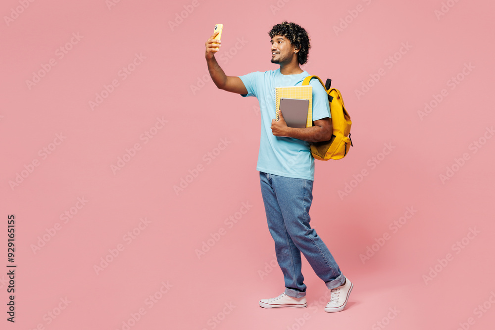 © ViDi Studio - Full body young Indian boy student in blue t-shirt casual clothes backpack bag hold books do selfie shot on mobile cell phone isolated on plain pink background High school university college concept