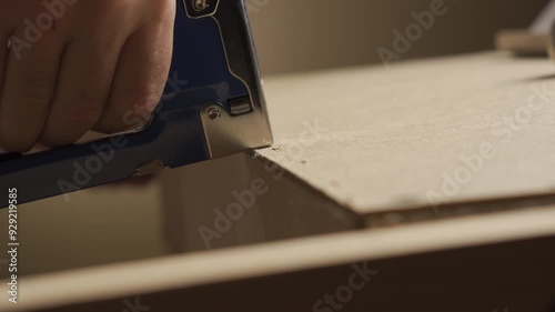 A man uses an industrial stapler to connect wooden parts while assembling furniture. Close-up of his hands
