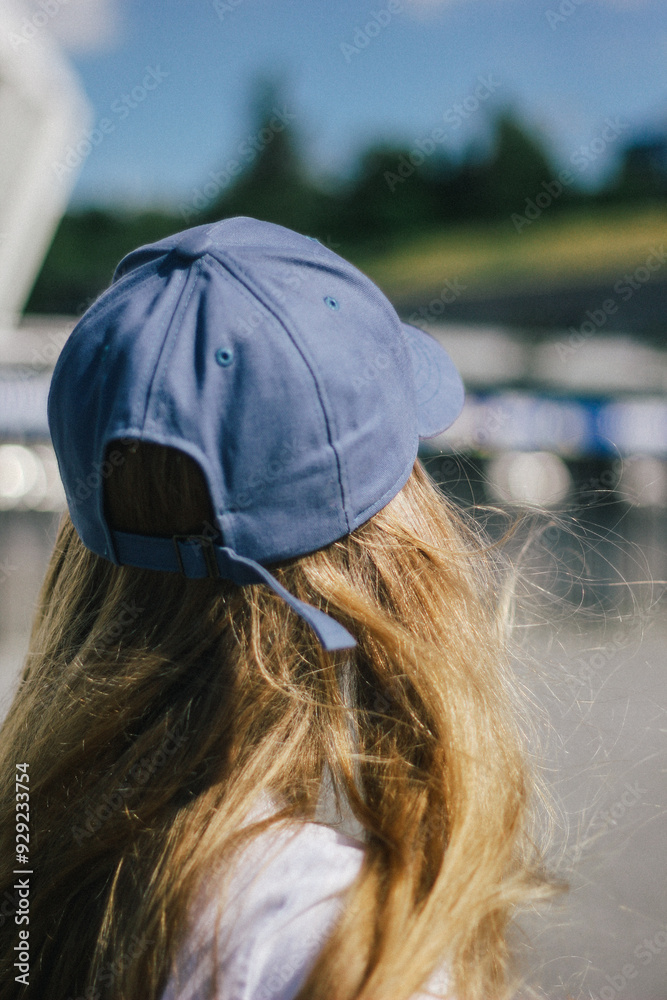 Back view of a young woman with long blonde hair, wearing a blue cap, looking out into the distance. The focus on her hair and the cap in natural light creates a serene, contemplative mood.