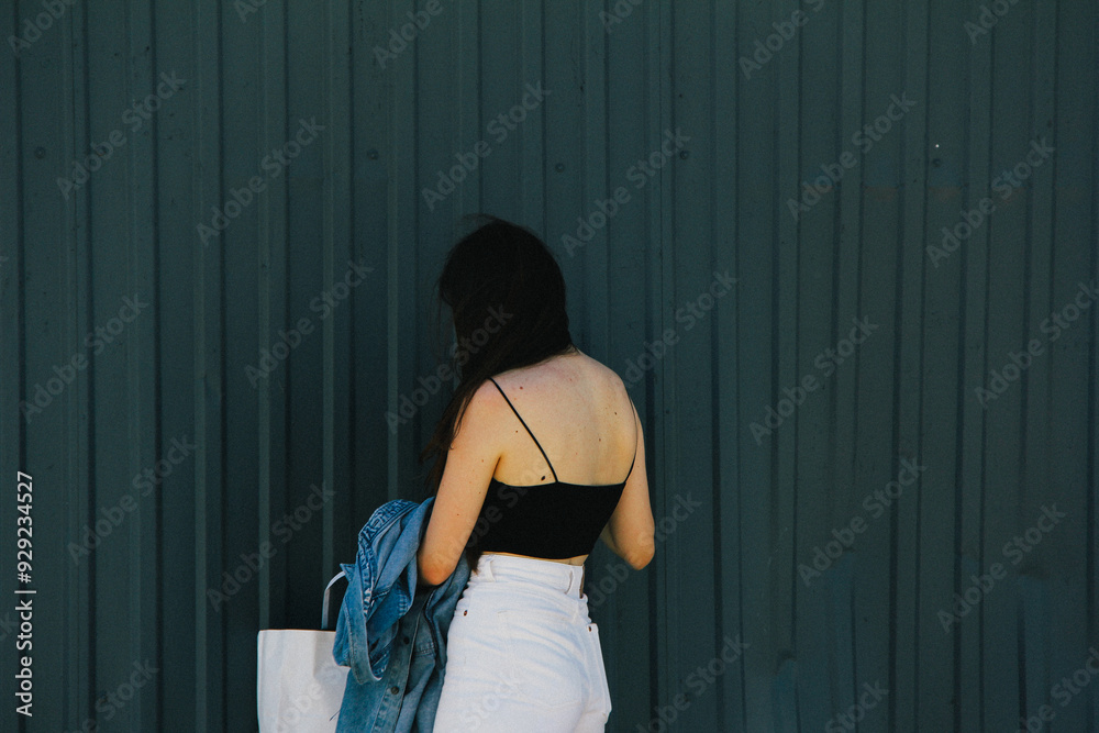 A woman in a black top and white high-waisted pants with her back to the camera, holding a denim jacket. The minimalist style emphasizes modern urban fashion.