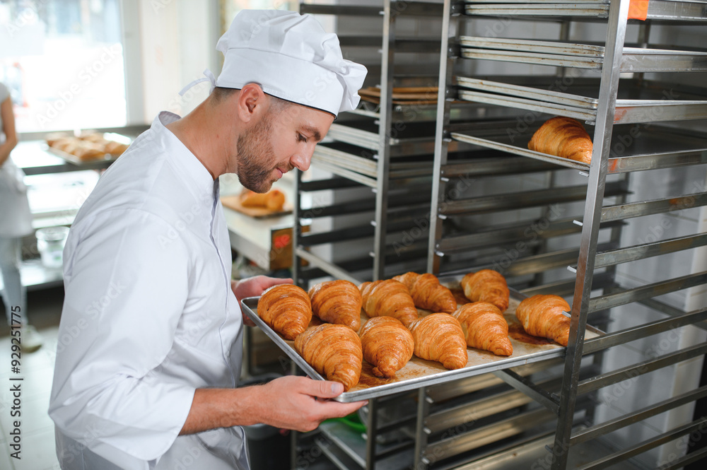 Handsome baker in uniform holding tray full of freshly baked croissants at the manufacturing Handsome baker in uniform holding tray full of freshly baked croissants at the manufacturing