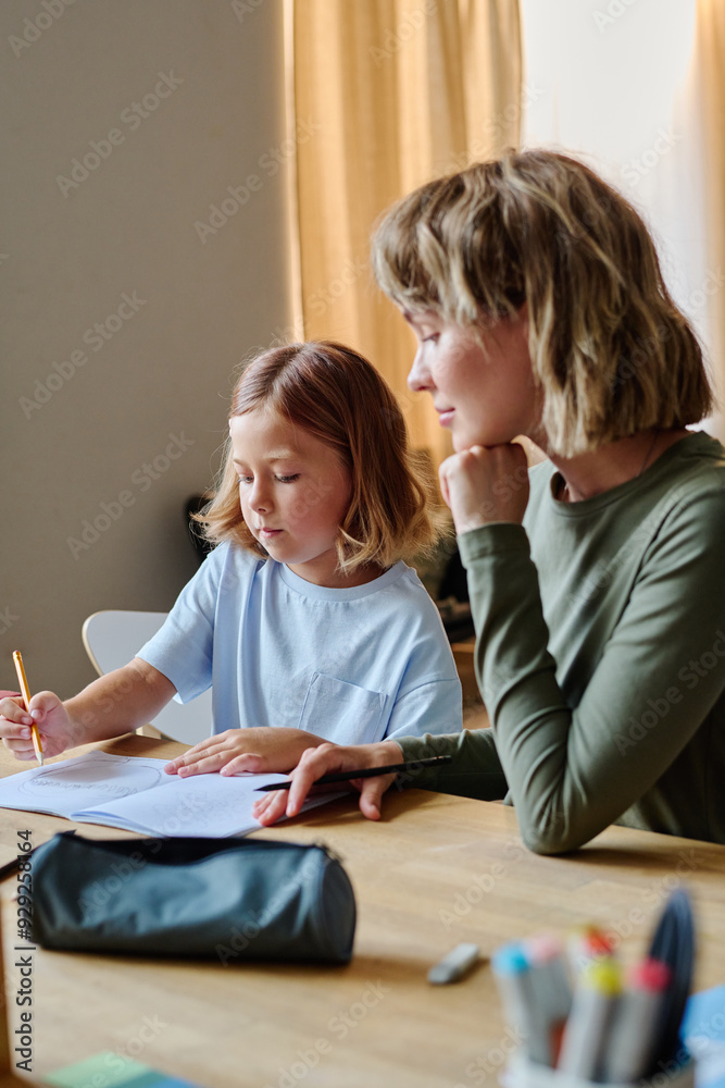 Fototapeta premium Young blonde mother sitting next to her lovely daughter while they doing homework