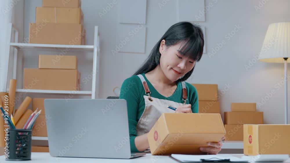 Asian sme businesswoman writing an address on a cardboard box while using a laptop in a home office setting Asian sme businesswoman writing an address on a cardboard box while using a laptop in a home office setting