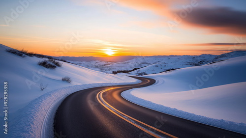 A picturesque winding road cutting through snow-covered hills as the sun sets, casting a golden hue over the serene winter landscape.