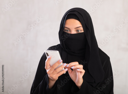 young light-skinned girl with her face covered by burka holds phone and types text message communicates with friends in interest chat