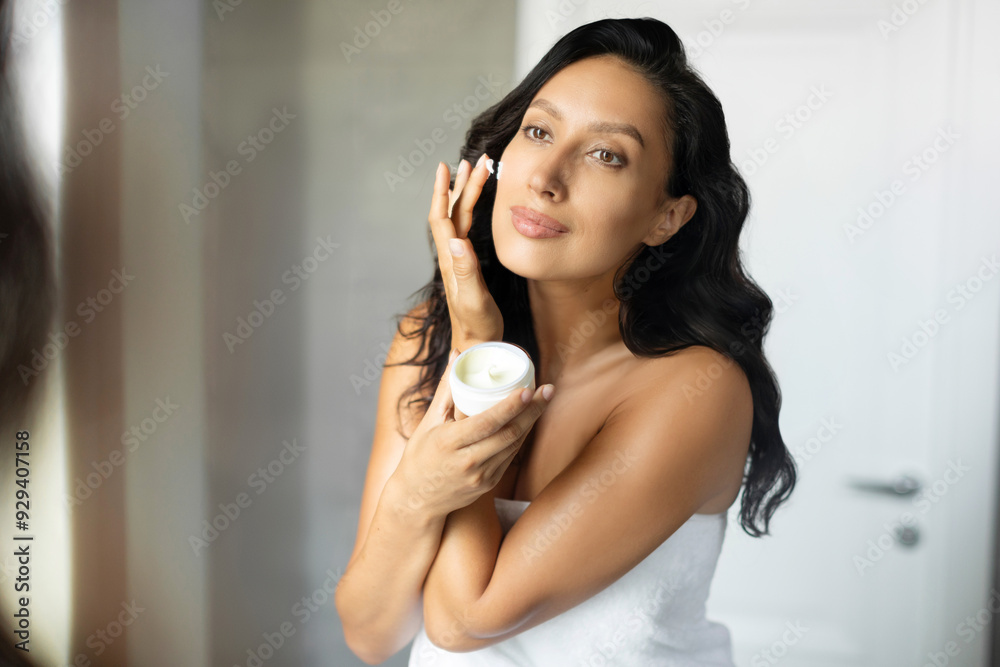 Woman caring of her facial skin, standing near mirror in bathroom, lady applying moisturizer, holding little jar of bio skin cream Woman caring of her facial skin, standing near mirror in bathroom, lady applying moisturizer, holding little jar of bio skin cream