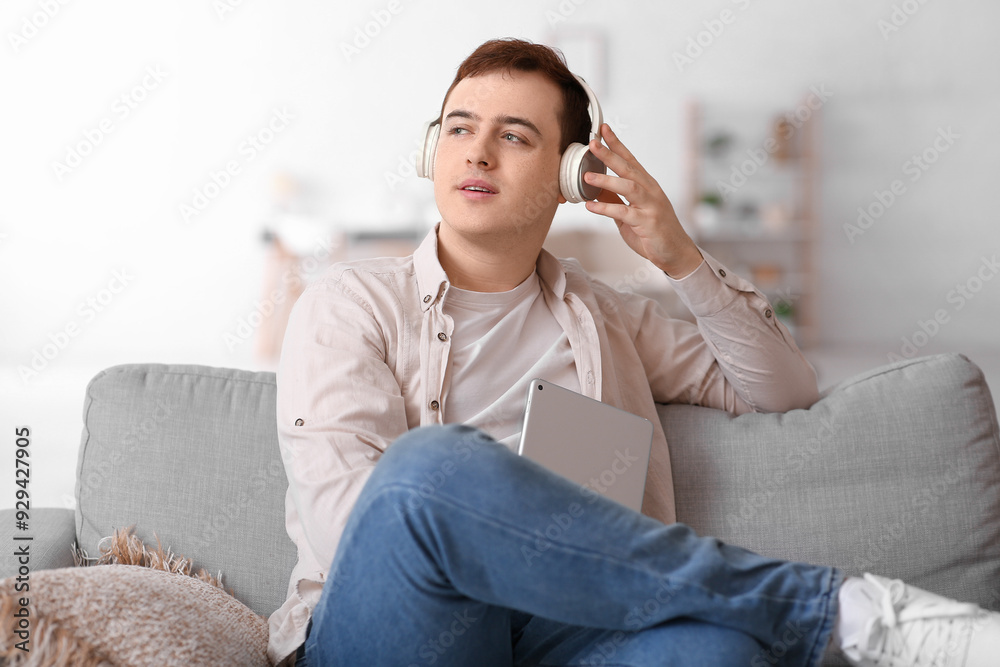Fototapeta premium Young man listening to audiobook and using tablet on sofa in living room