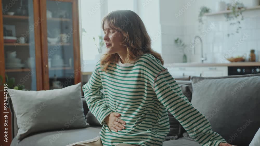 Young sick woman sitting on couch in living room clutching her stomach in pain, feeling burning sensation or cramps at home