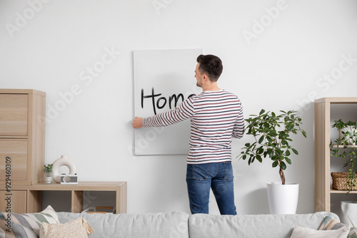 Young man hanging poster in light wall at home, back view