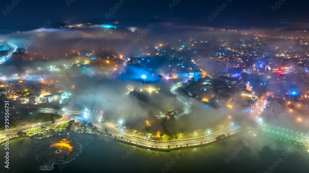 Fototapeta premium Aerial view of a Da Lat City in the mist magical night, hiden in the fog and clouds. Urban development texture, transport infrastructure. Tourist city in developed Vietnam. Night cityscape