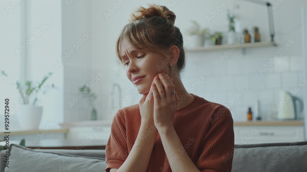 Young sick woman sitting on sofa during the day at home, touching her sore jaw and suffering from stubbing toothache Young sick woman sitting on sofa during the day at home, touching her sore jaw and suffering from stubbing toothache