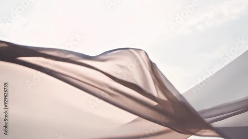 Light pink fabric gently blowing in the wind, creating elegant folds and curves against a serene sky background. The soft and airy nature of the fabric evokes a sense of grace and movement
