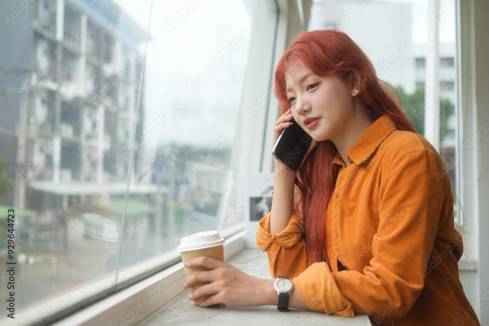 Fototapeta premium Attractive young woman with long red hair sitting by a large window, talking on mobile phone.