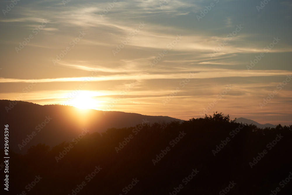 Sun sets behind rolling hills, casting warm, golden glow across sky. Wispy clouds, tinged with hues of orange and yellow, stretch across deepening blue sky. Serene and expansive landscape. Sun sets behind rolling hills, casting warm, golden glow across sky. Wispy clouds, tinged with hues of orange and yellow, stretch across deepening blue sky. Serene and expansive landscape.