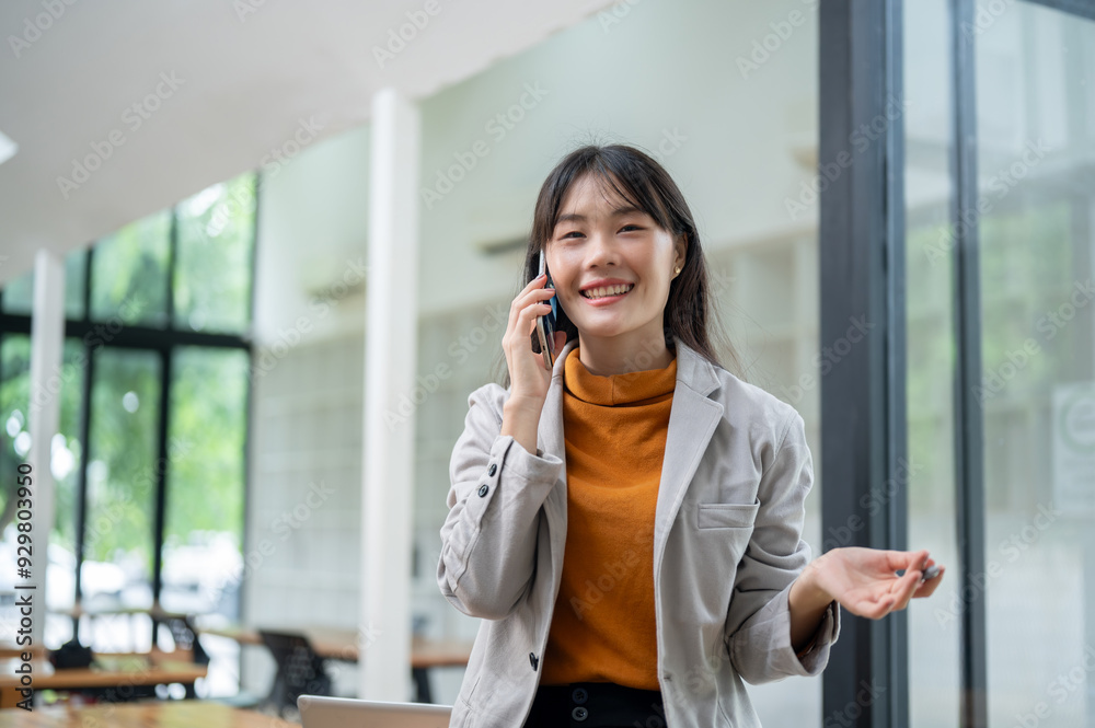 A confident Asian businesswoman is talking on the phone with her client while standing in the office A confident Asian businesswoman is talking on the phone with her client while standing in the office