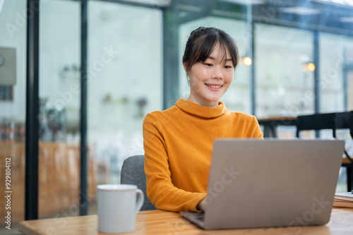 A charming Asian woman is working remotely from a coffee shop, working on her laptop computer.