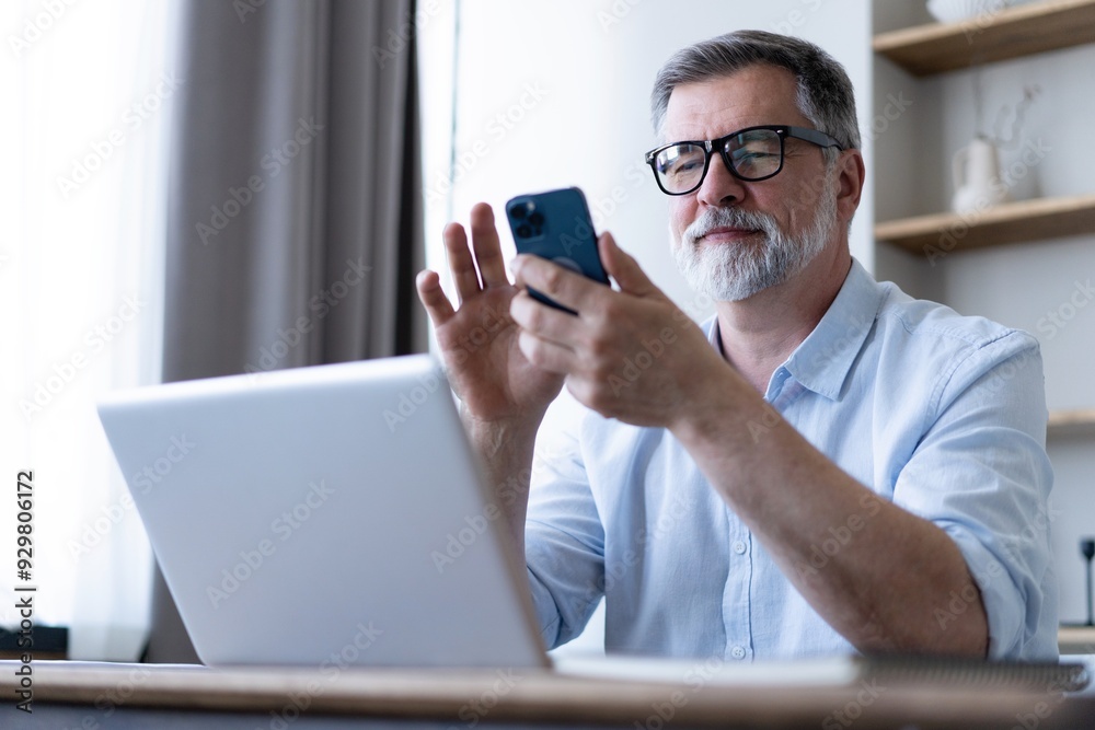 Handsome smiling senior man wearing glasses using mobile phone while sitting at his workplace with laptop at home Handsome smiling senior man wearing glasses using mobile phone while sitting at his workplace with laptop at home
